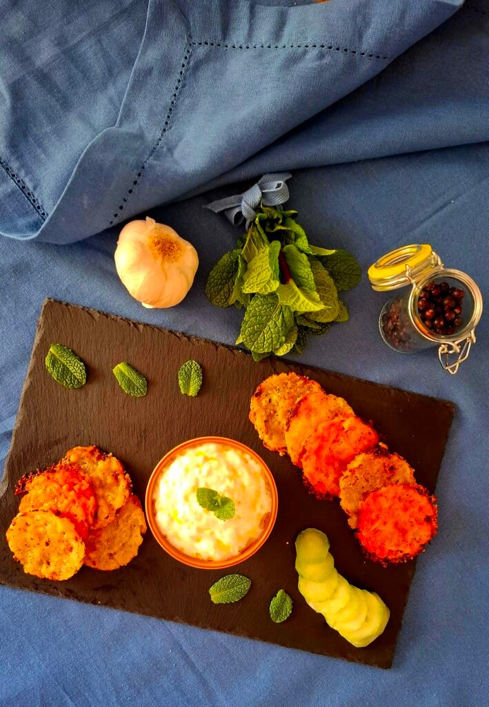 Cialde croccanti di carote e patate e parmigiano su piatto nero in pietra lavica con tovaglia blu a contrasto, accompagnate da salsa tzatziki 