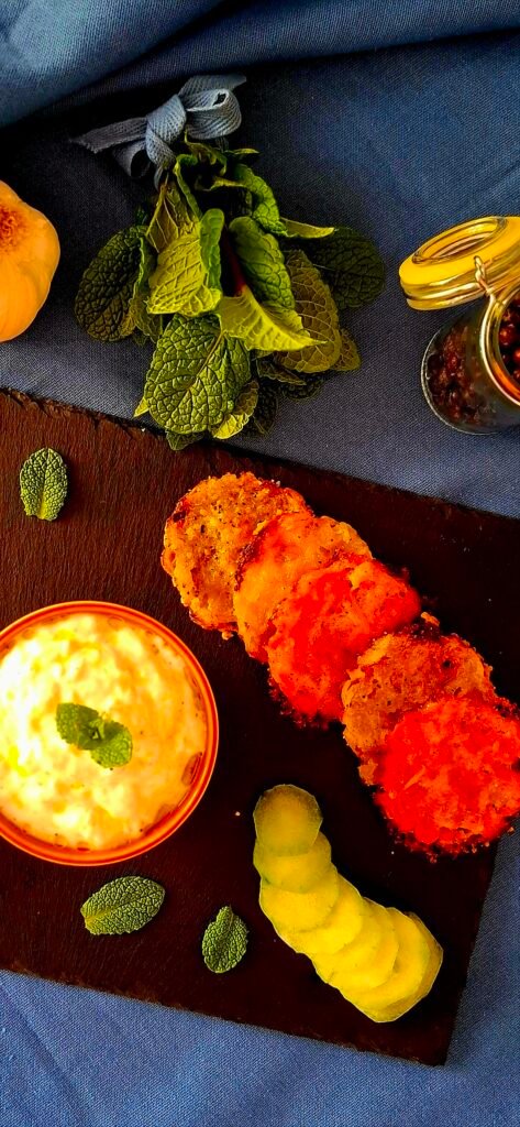 Cialde croccanti di patate e carote del bollito servite con salsa tzatziki e guarnite con menta fresca e un fiocchettino in tinta, presentate su una tovaglia a contrasto