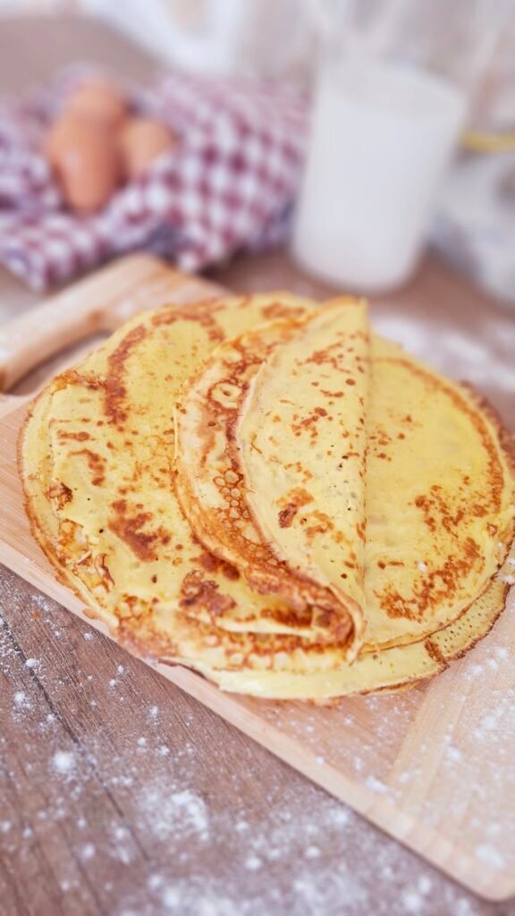 8 Crepes impilate una sopra l'altra su tagliere e sfondo con tovaglietta quadri latte e uova e farina