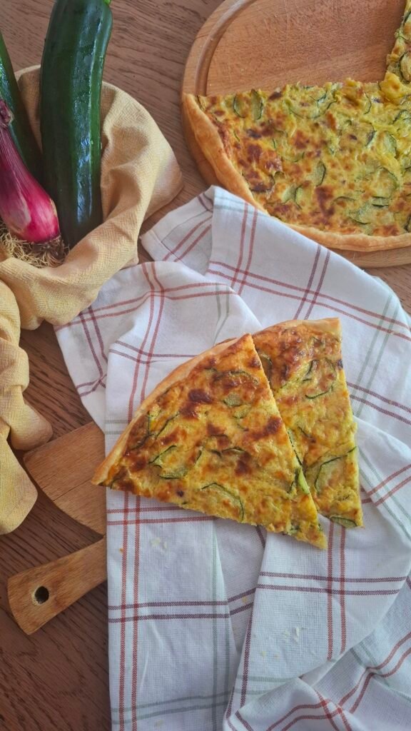 Fette di torta salata zucchine e crema di cipollotti su tovaglietta a quadri vicino a tagliere con torta quiche 