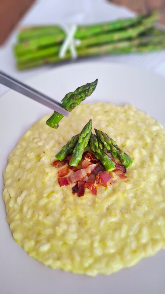 Pinza che adagia una punta di asparago sul risotto cremoso asparagi e pancetta croccante su tovaglietta di primavera e tavolo in legno