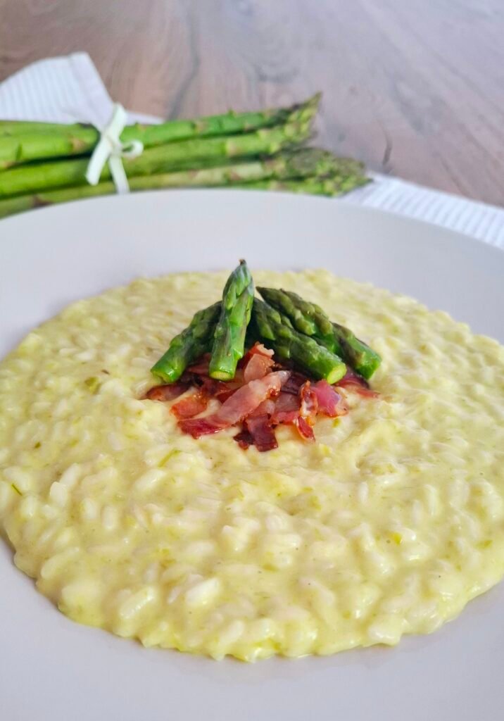 Risotto cremoso asparagi con pancetta affumicata croccante e punte di asparagi su piatto a contrasto bianco con tovaglietta primaverile 