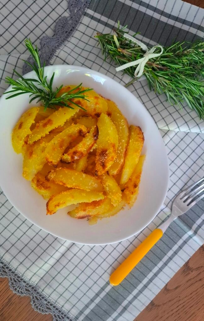 Spicchi di patate croccanti gratinati al forno su piatto bianco con rosmarino e tovaglietta grigia a contrasto