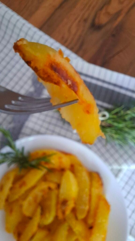 Primo piano patata croccante con forchetta,  sullo sfondo piatto con patate e rametto rosmarino con fiocchetto su tovaglietta bianca a quadri grigi che contrasta con giallo delle patate