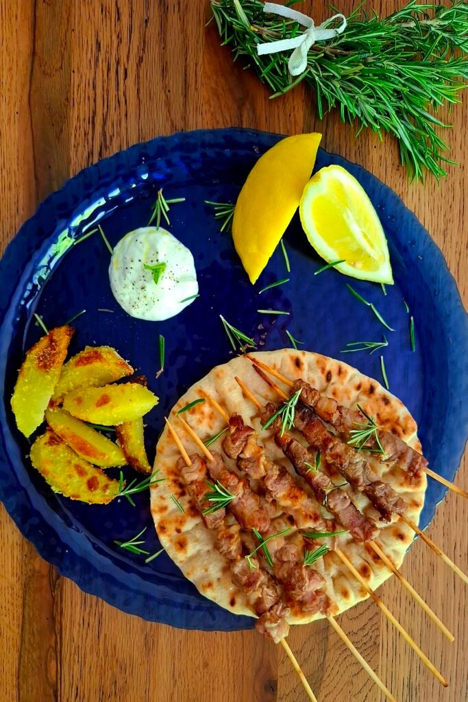 Spiedini maiale e arrosticini pecora adagiati su pane pita su piatto blu accompagnati con limone salsa tzatziki e patate croccanti al forno