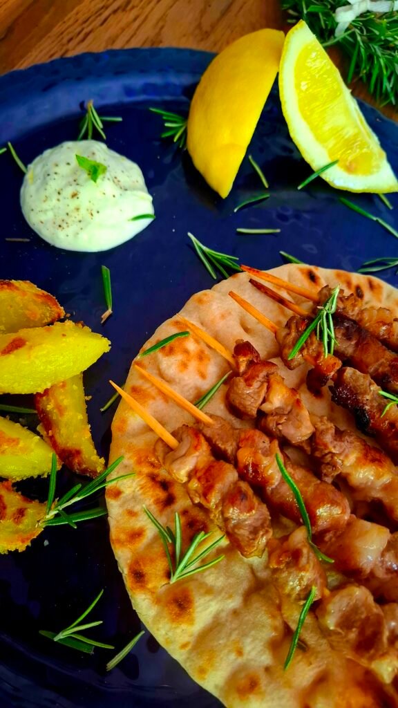 Spiedini maiale  e arrosticini pecora su pita su piatto blu a contrasto con contorno di patate al forno e tzatziki