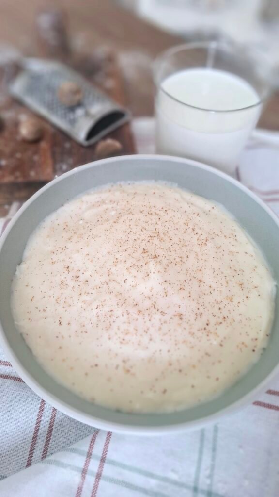 Besciamella cremosa senza grumi bianca perlato con contrasto di noce moscata in fondina a contrasto verde su tovaglietta a quadri con sfondo di grattugia con noce moscata e bicchiere latte