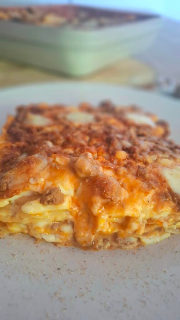Lasagna fetta con formaggio e sughetto che colano su piatto spolverato di noce moscata con sullo sfondo pirofila lasagne bolognesi cremose 