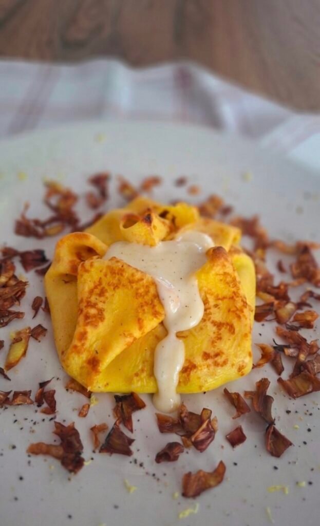 Rosa di crepes ai carciofi con besciamella alla fontina che cola sul piatto con attorno una corona di petali di chips di carciofo fritto croccante su tovaglietta a quadri verde e rosso e bianco 