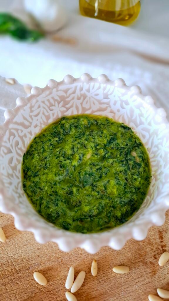 Pesto cremoso verde brillante in ciotola beige lavorata di bianco su tagliere legno con pinoli sparsi e oliera e basilico