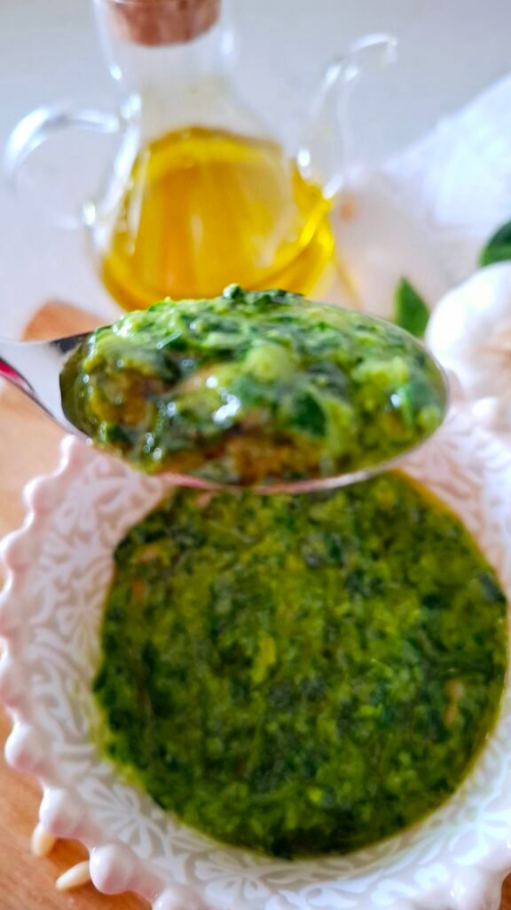 Zoom di Pesto sul cucchiaio color verde brillante dove si vede cremosità. Sullo sfondo Scodella con pesto e oliera