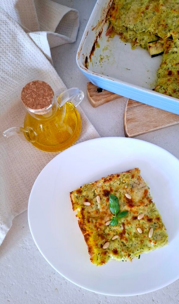 Fetta di lasagna pesto zucchine cotto scamorza in piatto bianco con pinoli olio e basilico come guarnizione .Sullo sfondo pirofila fumante di lasagne