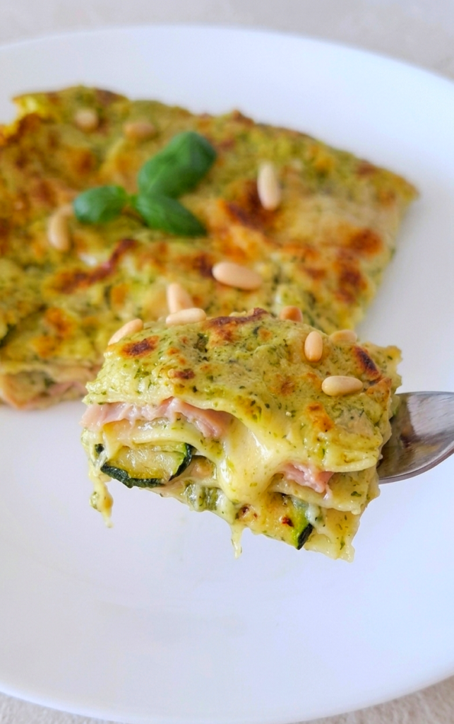 Boccone di lasagne filanti prese dalla forchetta. Lasagne di pesto di basilico prosciutto cotto zucchine e scamorza. Guarnite con basilico e pinoli.