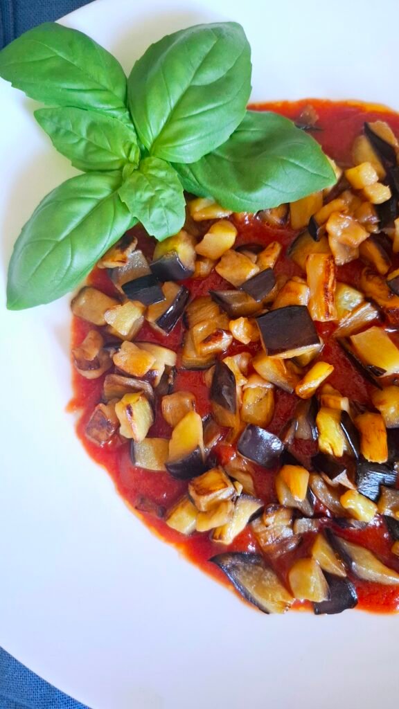 Melanzane a funghetto sopra salsa di pomodoro con basilico a forma di fiore su piatto bianco a contrasto. Si notano croccantezza delle melanzane e ricetta colorata.