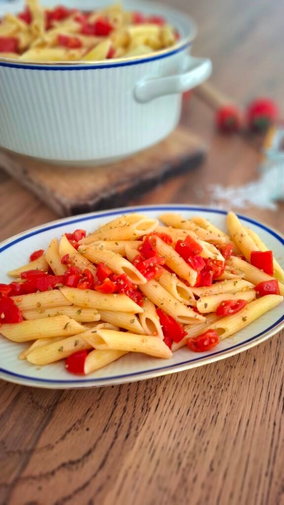 Piattino con pasta pomodorini origano aglio e olio e vicino terrina con pasta pomodorini su tagliere in legno e tavolo in legno con sfondo rametto di pomodorini e sale grosso che cade da vasetto in vetro