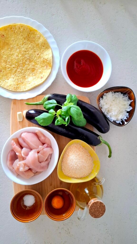 Ingredienti per tacos estivi sul tagliere: le tortillas, il petto di pollo a pezzetti, le melanzane, la passata di pomodoro, l'aglio, il peperoncino, l'uovo, il pane grattugiato, il basilico, l'olio extravergine d'oliva, il sale fino e il parmigiano a scaglie.