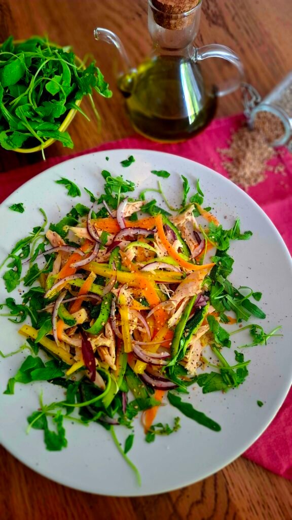 Insalata di pollo fredda molto colorata con pollo, carote, zucchine, peperoni, rucola, semi papavero e sesamo su piatto tondo chiaro a contrasto su tavolo legno con sfondo di rucola,oliera e semi caduti su tovagliolo rosa acceso