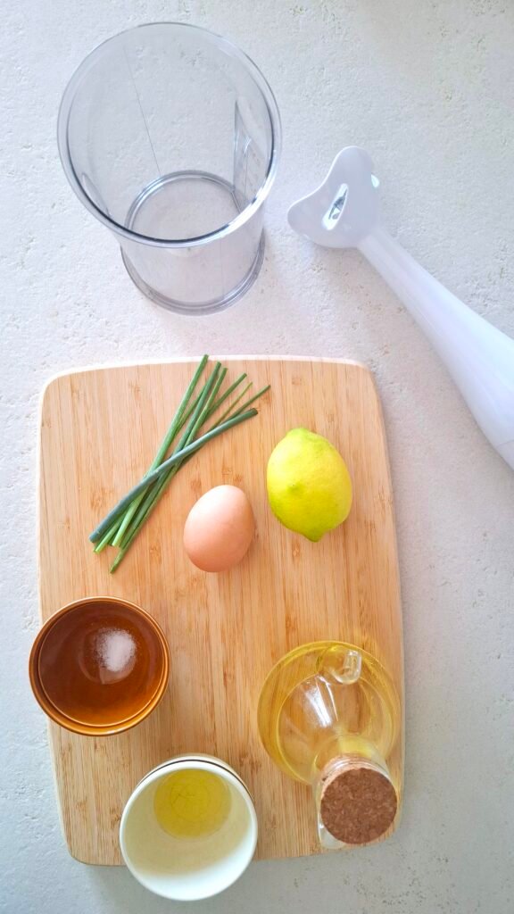 Tagliere con ingredienti maionese: l'uovo, il mix di olio di arachidi e di girasole, l'olio di oliva, il limone, l'erba cipollina e il sale fino.