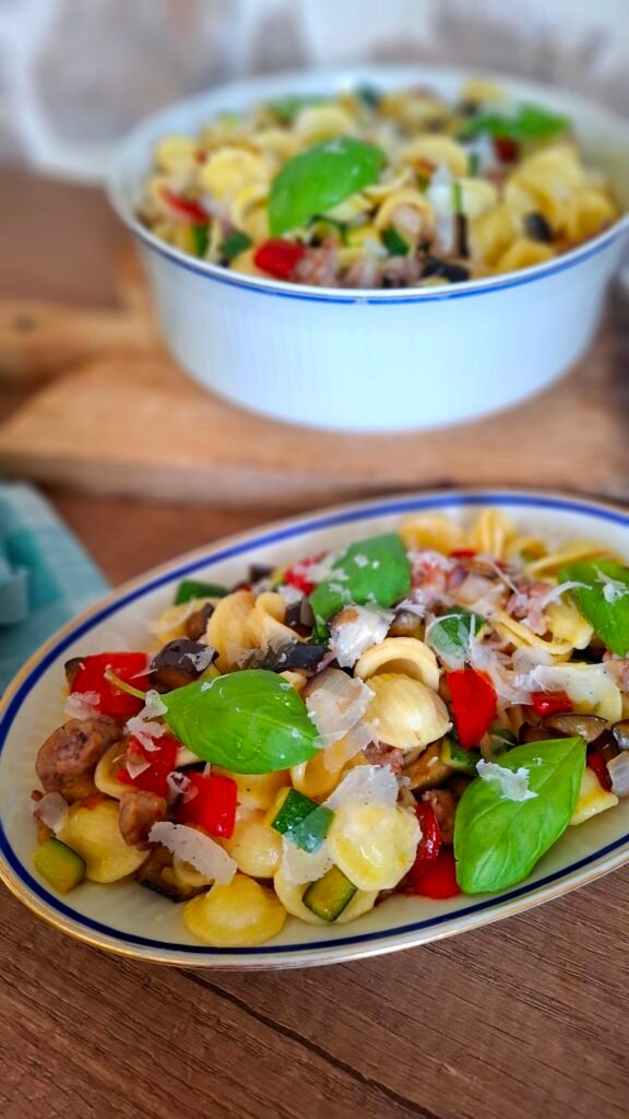 Pasta corta orecchiette con salsiccia e verdure km 0 cipolle, peperoni, melanzana, zucchine in un piatto ovale vicino a terrina colma di pasta salsiccia e verdure su tagliere legno con tovagliolo verde a quadri