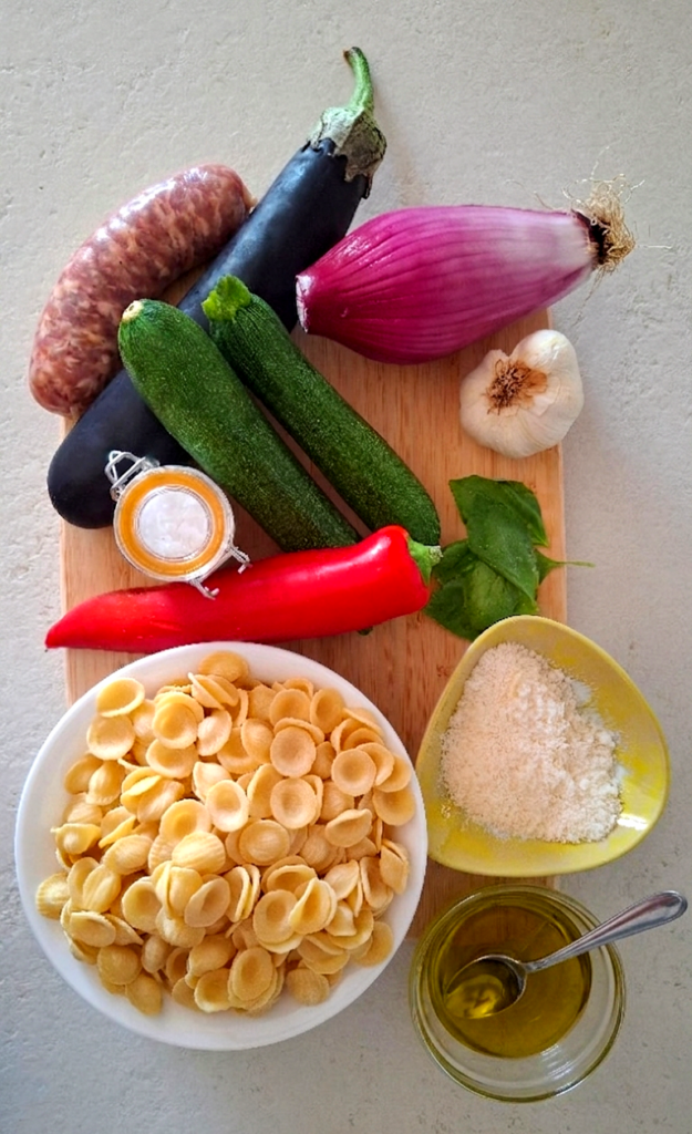  tagliere con ingredienti: le orecchiette, le melanzane, le zucchine, i peperoni, la mezza cipolla, l'aglio, il basilico fresco, l'olio extravergine d'oliva, il sale grosso e il formaggio grattugiato.