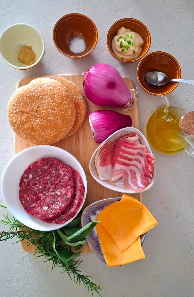 tutti gli ingredienti su tagliere in legno: i panini per l'hamburger, le svizzere di manzo, il cheddar, il bacon, la cipolla, il miele, la senape, la maionese all'erba cipollina, salvia e rosmarino, l'olio extravergine di oliva e il sale.