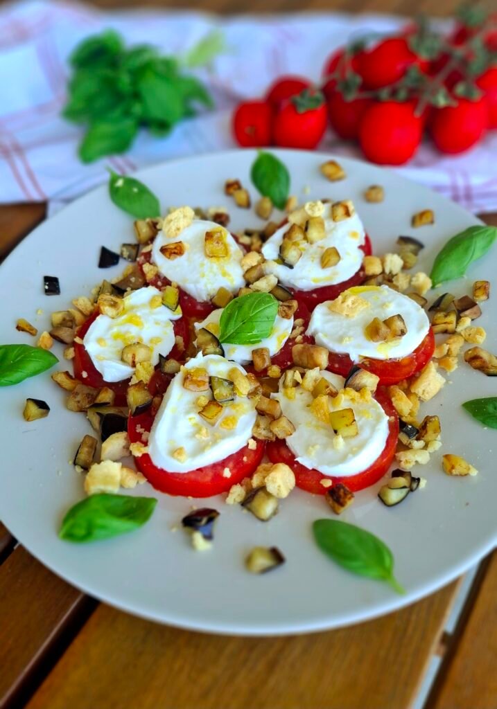 Piatto bianco tondo con parmigiana fredda di melanzane stile caprese aforma di fiore...pomodori, bufala, melanzane non fritte a cubetti e taralli sbriciolati . Sullo sfondo canovaccio con pomodori e rametto basilico.