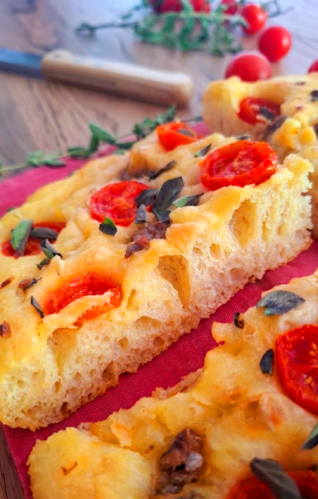 Focaccia mediterranea morbida con pomodorini alici crema aglio e origano fresco su tagliere legno e sfondo con rametti di origano