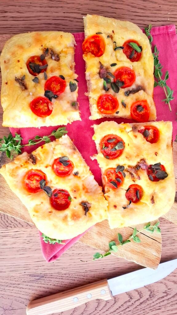 Focaccia mediterranea morbida e profumata con alici crema di aglio pomodorini origano fresco su tagliere legno con tovagliolo rosa e coltello in legno di fianco
