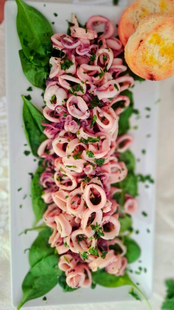 Insalata calamari su letto spinacini impiattata su piatto bianco lungo e stretto molto scenico: i calamari sono morbidi e ricchi di citronette e prezzemolo con bruschettine calde e olio.