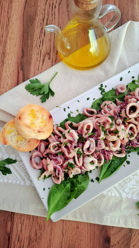 Insalata di calamari su letto di spinacini con bruschettine in piatto rettangolare lungo . È condita con citronette e prezzemolo. È morbida e succosa.