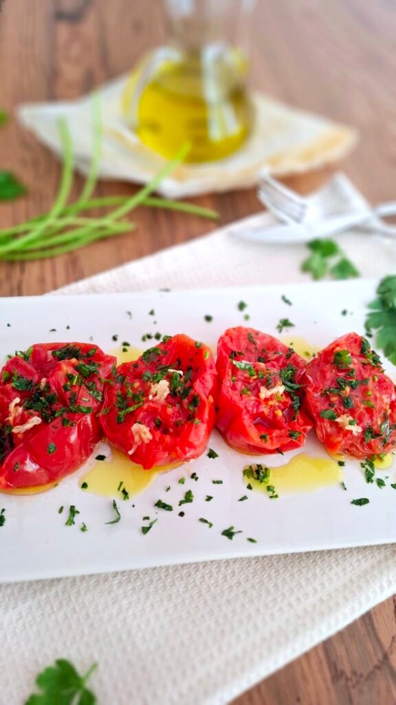Pomodori cotti in padella presentati in piatto lungo conditi con crema di aglio olio e prezzemolo. Su tavolo in legno con tovaglietta beige e posate eleganti