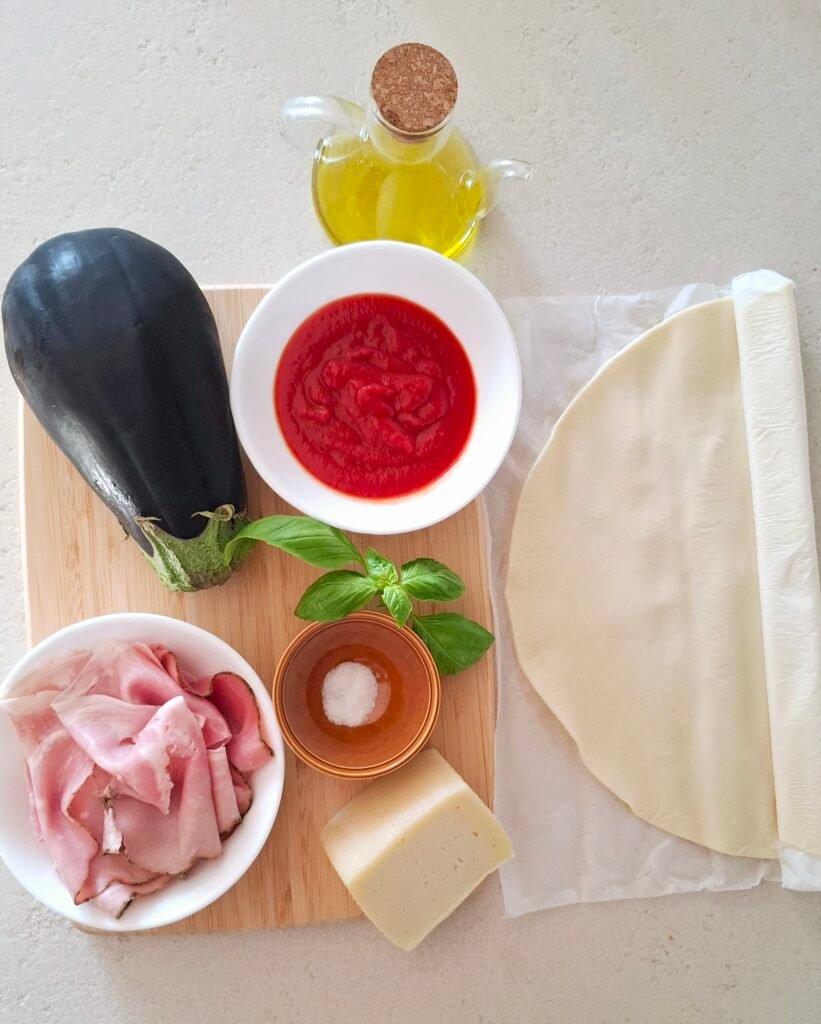 tutti gli ingredienti su tagliere: la pasta sfoglia, la melanzana, il prosciutto di Praga, la caciotta mista, la passata di pomodoro, il basilico, l'olio extravergine di oliva e il sale fino.