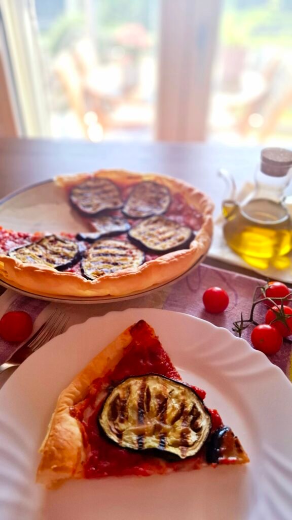 Torta salata parmigiana di melanzane e Praga affumicato e caciotta servita in piattino con a fianco olio e pomodori su tovaglietta lillà, sullo sfondo finestra che dà sul giardino di casa