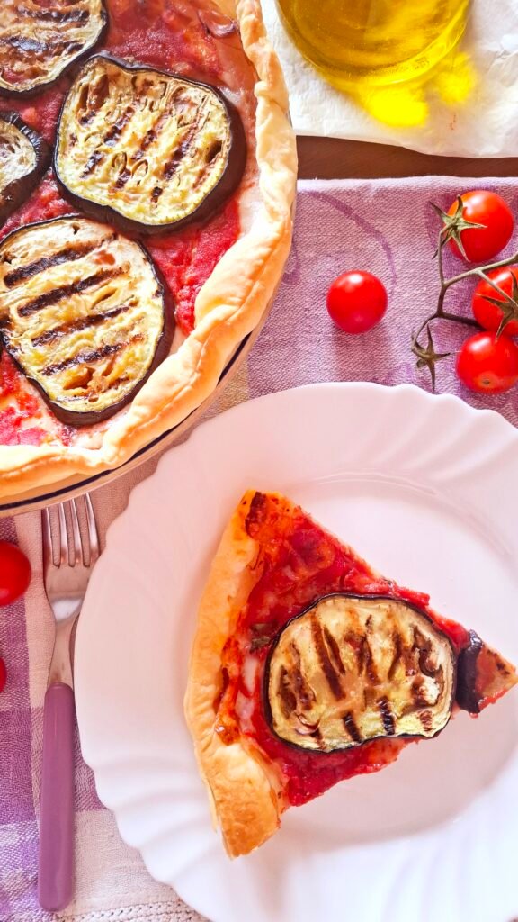Torta salata di melanzane su piatto bianco con fetta tagliata. Versione estiva e profumata della parmigiana servita su tovaglietta lilla con posate lilla e pomodorini sparsi sul tavolo
