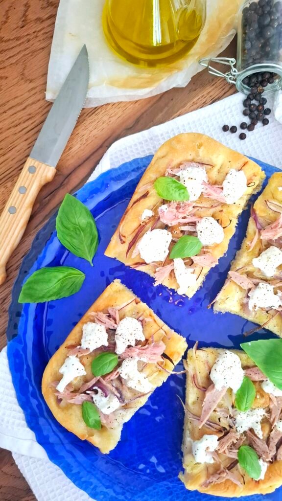 Focaccia alta idratazione gourmet con tonno, cipolla e burrata su vassoio blu a contrasto con basilico olio e pepe