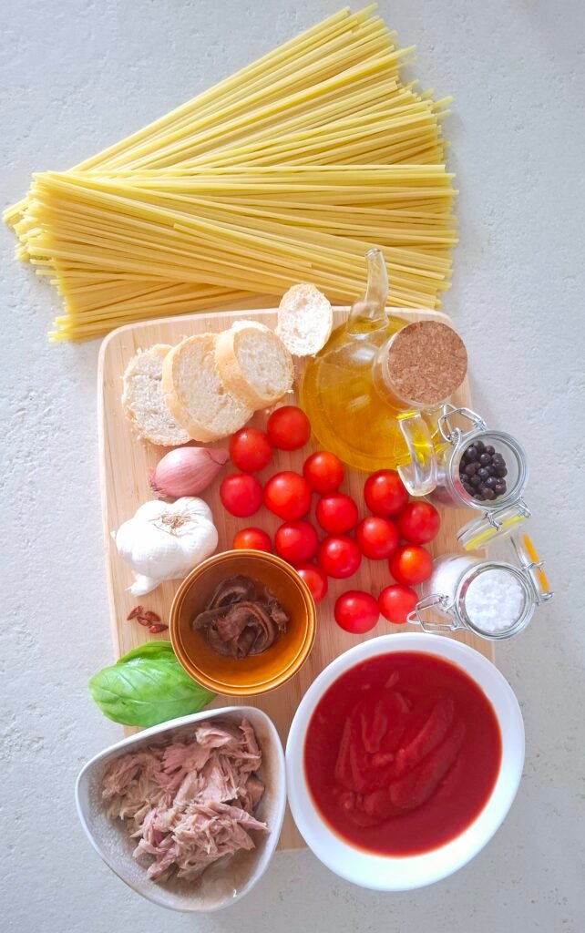 tutti gli ingredienti sul tagliere: le linguine, la passata di pomodoro, i pomodorini,il sale grosso, il tonno, le alici, le fette di pane, l'aglio, lo scalogno, il peperoncino, il pepe, l'olio evo e il basilico.