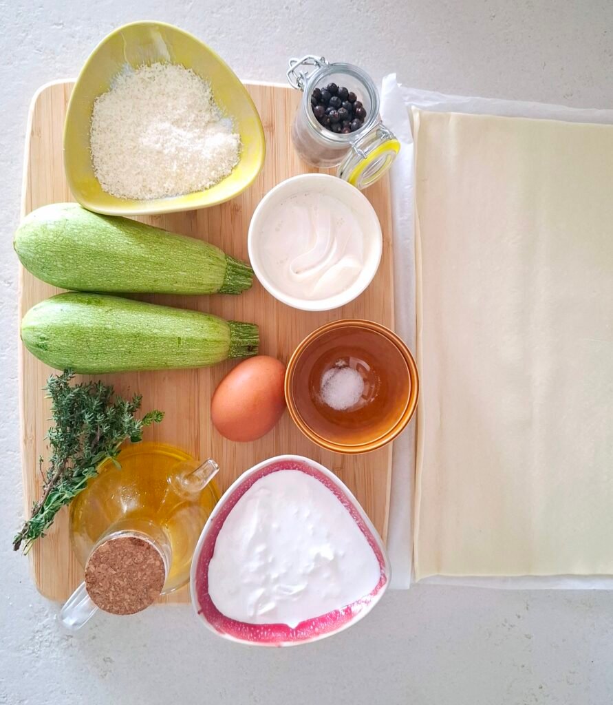 Ingredienti su piano di lavoro: pasta sfoglia rettangolare, le due zucchine, l'uovo, il parmigiano reggiano, la panna e la stracciatella, il timo, l'origano, il sale fino, il pepe e l'olio extravergine d'oliva.