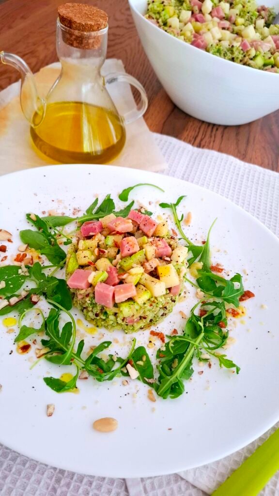 Insalata di riso integrale e quinoa presentata a forma di tortino con pesto rucola dadini di prosciutto Praga e asiago e zucchine su piatto bianco con sbriciolata di mandorle e pinoli 