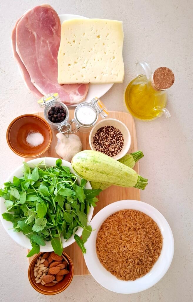 Ingredienti su tagliere: riso integrale, quinoa, Asiago, prosciutto di Praga, rucola, zucchine, mandorle, pinoli, olio extravergine d'oliva,aglio sale grosso, sale fino e pepe nero.