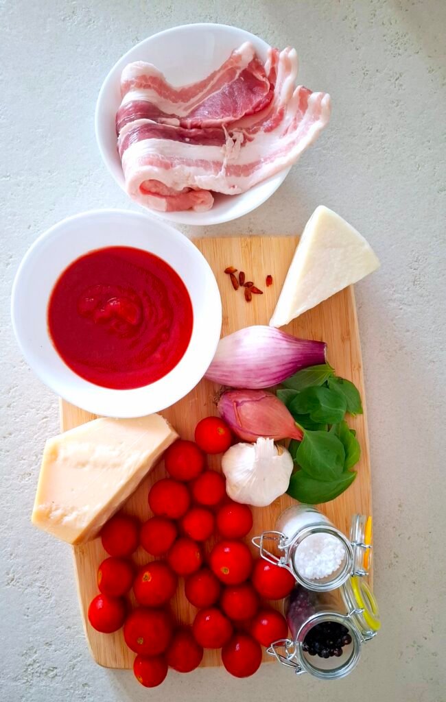 pancetta stesa, la passata di pomodoro, i pomodorini ciliegia, il pecorino romano, il parmigiano, il basilico, il peperoncino, il pepe nero, il sale grosso, la cipolla, l'aglio e lo scalogno su tagliere