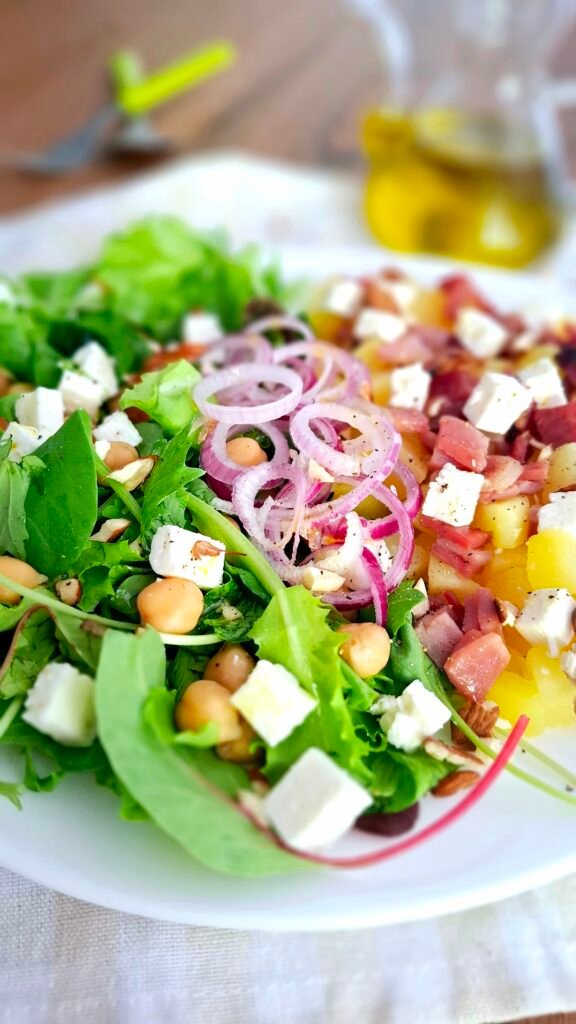 Piatto ovale bianco con insalatona di misticanza con feta, patate, speck, ceci e cipolla tropea su tovaglietta quadri beige e sfondo di oliera posate verde acido e mandorle sbriciolate