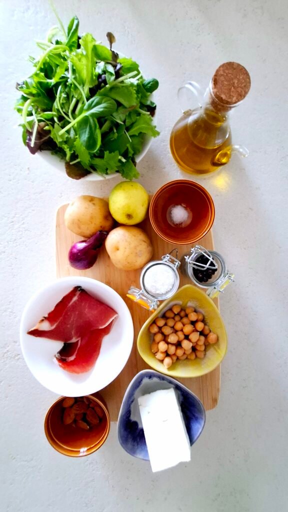 Sul tagliere: misticanza, patate novelle, feta, speck, ceci precotti, olio extravergine d'oliva, mandorle, cipollotto, sale grosso, sale fino, pepe e succo di limone