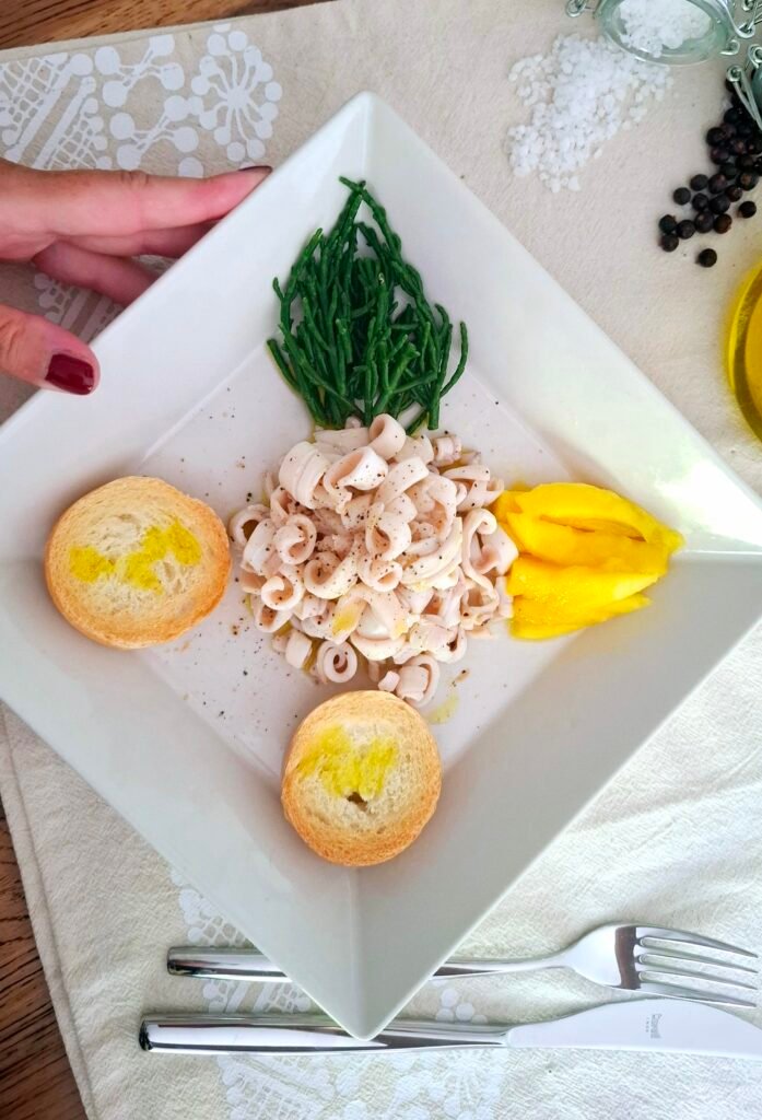 Mia mano che tiene il piatto quadrato con calamari salicornia mango e bruschettine su tovaglietta beige e posate eleganti