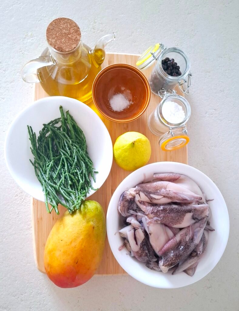 sul piano di lavoro tutti gli ingredienti necessari: i calamari, la salicornia, il mango, il limone, l'olio extravergine d'oliva, il pepe, il sale grosso e il sale fino.