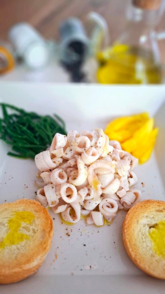 Insalata calamari morbidi bolliti con salicornia mango e bruschette su piatto quadrato con sfondo di oliera e spezie