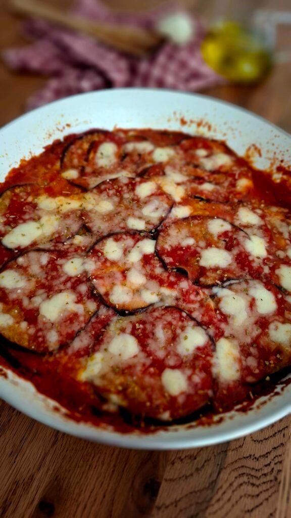 Parmigiana melanzane filanti e morbide con scamorza affumicata origano in padella bianca su tavolo legno con cucchiaio legno e tovagliolo quadretti rossi