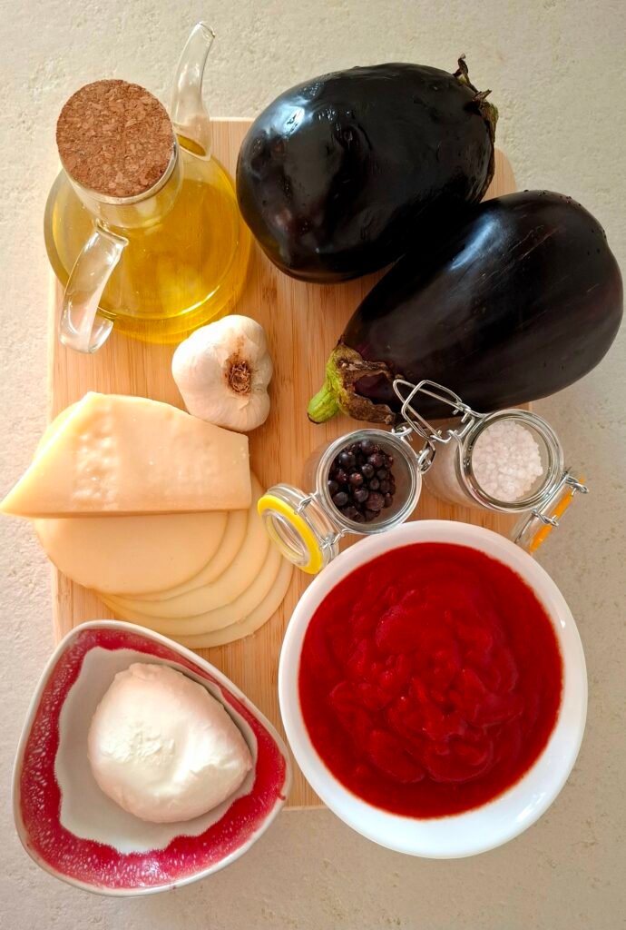 Sul tagliere ingredienti per parmigiana: melanzane, olio evo, passata di pomodoro, aglio, pepe, sale grosso, sale fino, mozzarella, scamorza e parmigiano reggiano