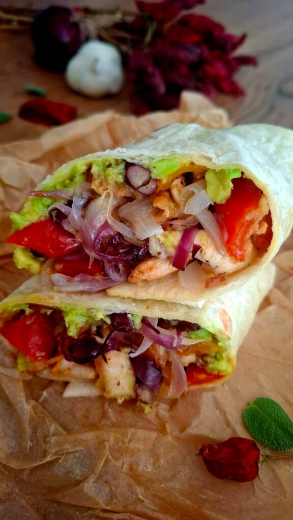 Burrito diviso in 2 disposto su carta bene da forneria con zoom su ripieno succoso di cipolle fagioli peperoni pollo bacon guacamole . Sullo sfondo peperoncino secco aglio salvia e cipollotto