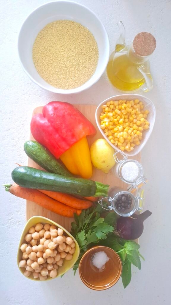 Sul tagliere ingredienti per couscous verdure: Couscous, peperone, zucchina, carota, mais, ceci, cipollotto, limone, prezzemolo, basilico, olio, pepe, sale grosso e sale fino.