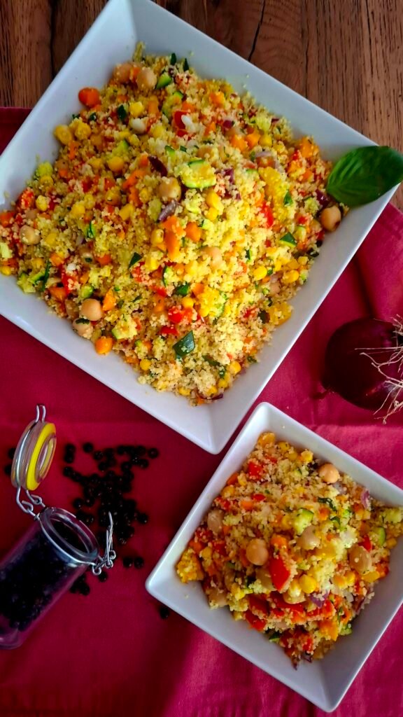 Cous cous di verdure coloratissimo servito in coppetta quadrata e rispettivo recipiente grande quadrato su tovagliolo rosa. Ci sono carote Zucchine peperoni mais cipollotto e ceci invitanti e croccanti